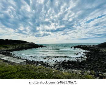 A pebble rocky coast with waves crashing against the shore. The sky is blue with white clouds, and there are distant mountains. - Powered by Shutterstock