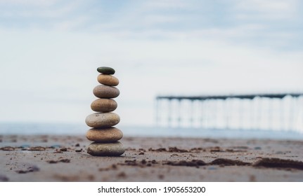 Pebble tower by the seaside with blurry pier down to the sea, Stack of zen rock stones on the sand, Stones pyramid on the beach symbolizing, stability, harmony, balance with shallow depth of field.