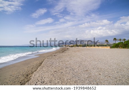 Pebbly beach in Motril Spain with azure coastal waters. Wide beach of southern Spain with mountains in a distance, clear water and partly cloudy blue sky. 