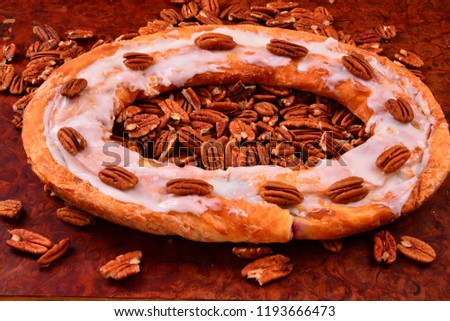Pecan Kringle oval with pecans on top and laying on a pile of fresh pecans on a burl surface. 