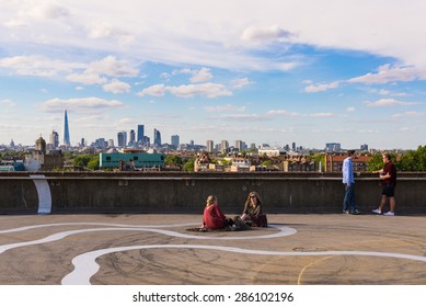 Peckham, London, UK - June 06 2015: Two Girls And Two Guys Drinking Beer On The Floor Of A Rooftop Bar Cafe In South-east London.View Of The City Of London Skyscrapers And The Shard In The Background.