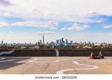 Peckham, London, UK - June 06 2015: Two Girls Sit On The Floor Of A Rooftop Bar Cafe In South-east London.View Of The City Of London Skyscrapers And The Shard In The Background.