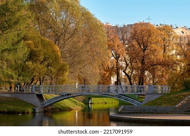 Pedestrian Bridge Across The Canal In The City Autumn Park, No People. Trees In Colorful Autumn Foliage Next To A Pond.