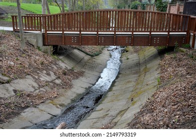 pedestrian bridge over a small gorge and a stream. consists of two steel crossbars. because  surface and railings of the bridge form brown painted planks. glued beams to the arch, concrete creek bed
