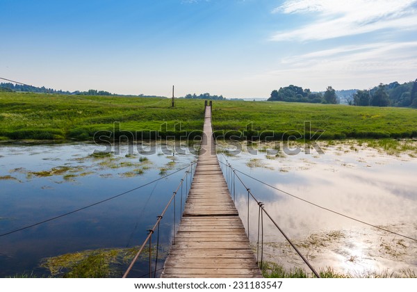 Pedestrian suspension bridge of steel and wood\
over the river, summer in\
Russia