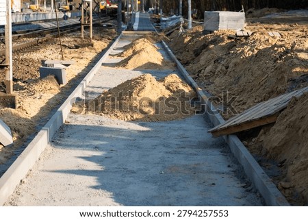 A pedestrian walkway, platform, or pavement under construction with concrete curbs, piles of sand, and a railway track running parallel to it.