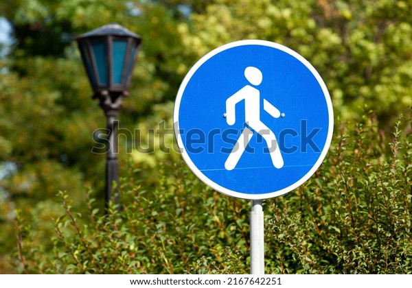 pedestrian zone\
road sign close-up on a background of park green trees. round blue\
road sign with white human icon. pedestrian zone in the park road\
sign next to a vintage street\
lamp.