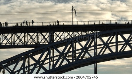 Pedestrians walking along the upper deck of the Dom Luis I Bridge, creating a silhouette against the cloudy sky while the intricate steel structure forms a graphic pattern below. Urban infrastructure