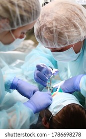 Pediatric Dentist And Assistant During Surgery Under General Anesthesia. Removal Of Deep Caries In A Child. Copy Space. Vertical Photo. People Are Out Of Focus.