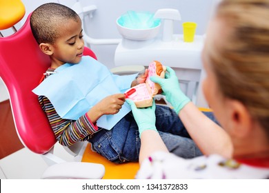 a pediatric dentist teaches an African American child who sits in a dental chair to brush his teeth properly. Pediatric dentistry