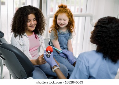 Pediatric dentistry. Back view of African lady dentist, showing how to brush teeth on jaw model, to her little patients, two smiling multiracial shcool girls, African American and Caucasian
