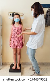 The Pediatric Doctor Checks The Caucasian Child And They Are Protected By A Face Mask Against The Coronavirus. Check Her Weight And Height On The Clinic Scale. The Nurse Wears Her White Coat.