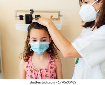 The pediatric doctor checks the Caucasian child and they are protected by a face mask against the coronavirus. Check her weight and height on the clinic scale. The nurse wears her white coat.
