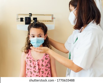 The Pediatric Doctor Checks The Caucasian Child And They Are Protected By A Face Mask Against The Coronavirus. Check Her Weight And Height On The Clinic Scale. The Nurse Wears Her White Coat.