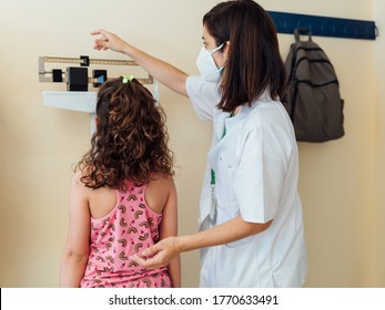 The Pediatric Doctor Checks The Caucasian Child And They Are Protected By A Face Mask Against The Coronavirus. Check Her Weight And Height On The Clinic Scale. The Nurse Wears Her White Coat.