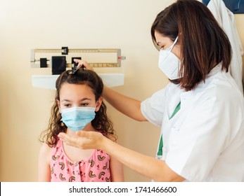 The pediatric doctor measures and weighs the child in the clinic office. They both wear a mask to protect against the covid-19 virus. The young girl is examined in the hospital to see how she is growi