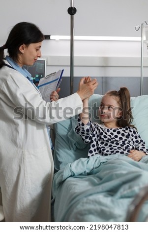 Pediatric expert doing high five gesture with ill kid sitting in patient bed. Pediatrician high fiving sick little girl under treatment resting in children healthcare facility.