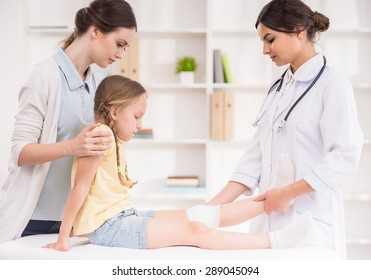 Pediatrician Doctor Bandaging Child's Leg. Mother Holding Baby In Her Hands.