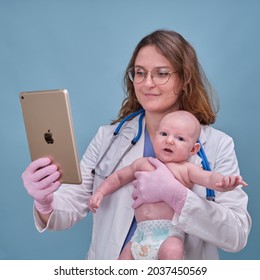 Pediatrician Doctor Holding Apple Ipad And Newborn Baby, Blue Studio Background. Happy Nurse In Uniform With Baby And Digital Tablet - Moscow, Russia, August 12, 2021