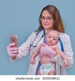 Pediatrician Doctor Holding IPhone Apple And Newborn Baby, Blue Studio Background. Happy Nurse In Uniform With A Child And A Mobile Phone - Moscow, Russia, August 12, 2021