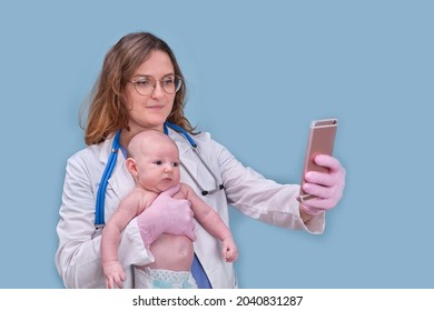 Pediatrician Doctor Holding Phone And Newborn Baby, Blue Studio Background. Happy Nurse In Uniform With A Child And A Mobile Phone