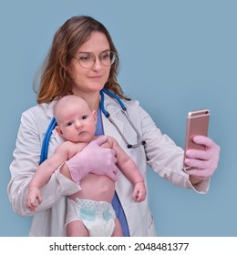 Pediatrician Doctor Holding Phone And Newborn Baby, Blue Studio Background. Happy Nurse In Uniform With A Child And A Mobile Phone