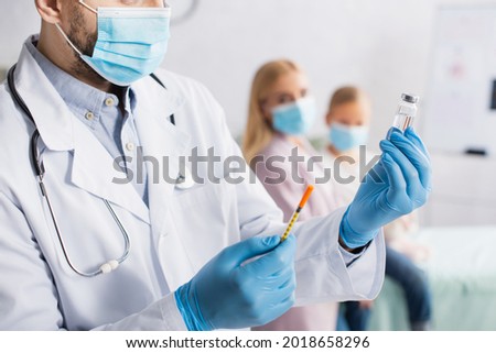 Pediatrician in latex gloves and medical mask holding vaccine and syringe near family on blurred background