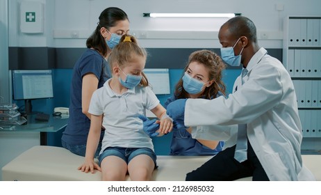 Pediatrician putting bandaid on injured arm of child at consultation in cabinet. Male doctor examining fracture to help little girl with recovery at medical appointment during pandemic.