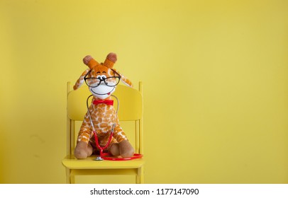 Pediatrician toy animal with stethoscope isolated on yellow background. Stuffed toy animal giraffe as pediatrician with stethoscope sitting on the chair with copy space. 