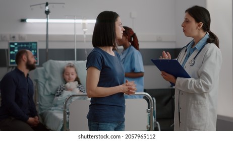 Pediatrician Woman Doctor Explaining Recovery Treatment To Worried Mother While Black Nurse Monitoring Sickness Symptoms. Sick Child Resting In Bed Recovering After Disease Breathing Surgery