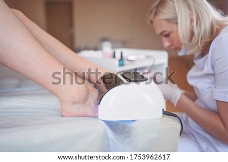 Pedicurist doing white nail polish on client legs using shellac lamp. Professional medical pedicure procedure. Foot treatment in SPA salon. Podiatry clinic. Beautician doctor hands in white gloves