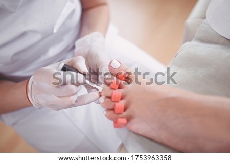 Pedicurist doing white nail polish on client legs using toe finger separator. Professional medical pedicure procedure. Foot treatment in SPA salon. Podiatry clinic. Beautician hands in white gloves