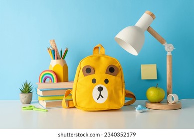 Peek into learning: side view photo of desk with pencils stand, pens, scissors, ruler, sharpener, lamp, plasticine, stack of books, plant, teddy-bear schoolbag, apple, set against blue wall backdrop
