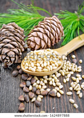 Peeled kernels of cedar nuts in a spoon, pine cones, green branches and nuts in the shell on a wooden board background