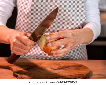 peeling onion by hand with a knife, home cooking, knife in a woman's hand, wooden board, onion peels, front photo. Checkered kitchen apron, white clothes