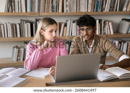 Peer collaboration. Two diverse multiethnic gen z students mates work at library desk use notebook paper sources to create joint scientific project discuss problem solution prepare for exams together