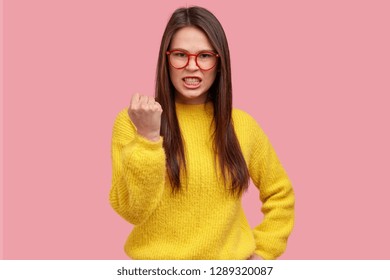 Peevish youngster shows fist with anger, clenches teeth, fights against injustice and violence, dressed in casual clothes, wears transparent glasses models against pink background, has determined look