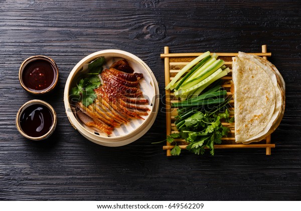 Peking Duck in bamboo\
steamer served with fresh cucumber, green onions, cilantro and\
roasted wheaten chinese pancakes with sauce Hoysin on black burned\
wooden background