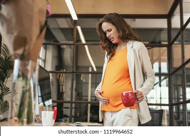 Pelvic discomfort. Low angle of beautiful pregnant CEO holding cup and touching belly