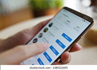 Penang, Malaysia - April 3, 2019: Woman Holding A Smartphone With Instagram Application On The Screen. Hand Touch On Follow Button. Blurred Cafe Interior On A Background.     