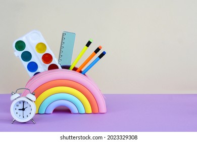 pencil case in the form of a bright rainbow with paints, pencils and a ruler and an alarm clock on a yellow-lilac background