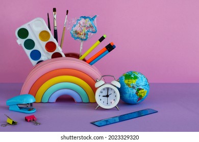 a pencil case in the form of a bright rainbow with a unicorn-shaped lollipop, paints, brushes, pencils, a white alarm clock, a stapler, a ruler and a toy planet on a lilac background