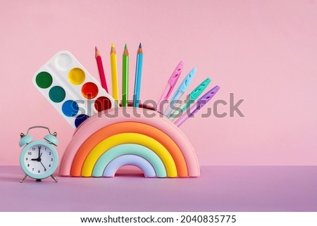pencil case in the form of a bright rainbow with paints, pencils and a ruler and an alarm clock on a pink background