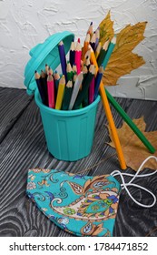 A pencil case in the form of a trash can. It contains colored pencils. Nearby is a protective face mask for the period of a pandemic. Dried maple leaves are added to the compositions.