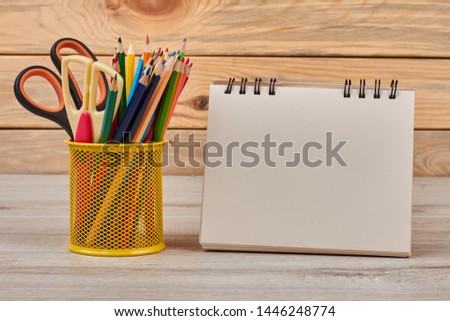 Pencils in metal basket and blank notebook. Set of colored pencils with scissors in metal holder and empty paper notepad. Education concept.