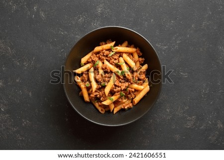 Penne pasta with minced meat, tomato sauce and greens in black ceramic bowl over dark stone background with copy space. Bowl of pasta bolognese. Top view, flat lay