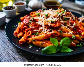 Penne mit Würstchen, Tomatensauce, Parmesan-Käse, Basilikum und Gemüse auf Holztisch
