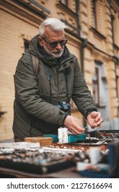 A Pensioner Is Looking For Rare Goods At A Flea Market. A Tourist Buys Vintage And Rare Jewelry From Pitchman.