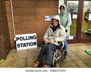 Pensioner in a manual wheelchair being pushed outside a polling station in the rain, during a General Election, UK