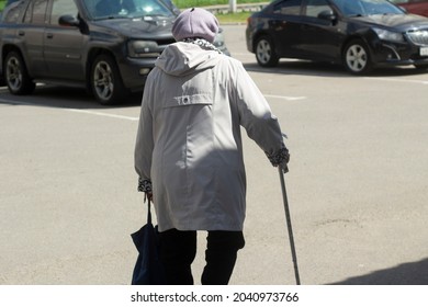 A Pensioner With A Stick Is Walking Along The Road. An Elderly Woman With A Sore Leg Leans On A Cane. Grandma Goes To The Store.